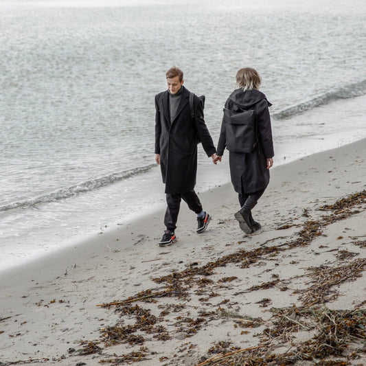 Man och kvinna går på en stranden en mulen dag med minimalistisk estetiskt vattentålig ryggsäck i polyester