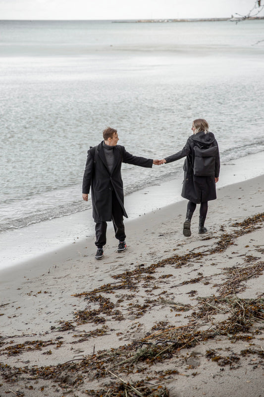 Man och kvinna håller handen på en strand. De har båda på sig en minimalistisk estetiskt vattentålig ryggsäck i polyester